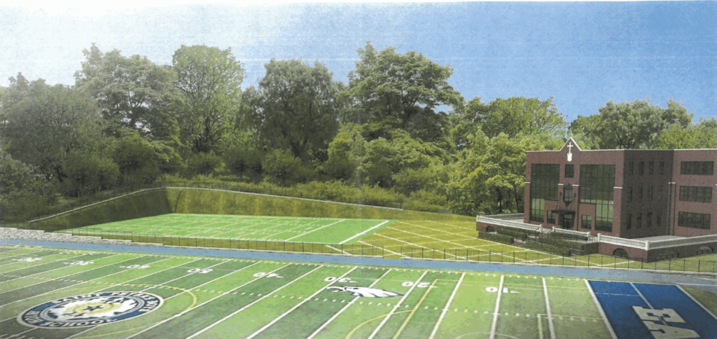 Football field and practice field beside a three-story brick school building, with dense trees in the background.