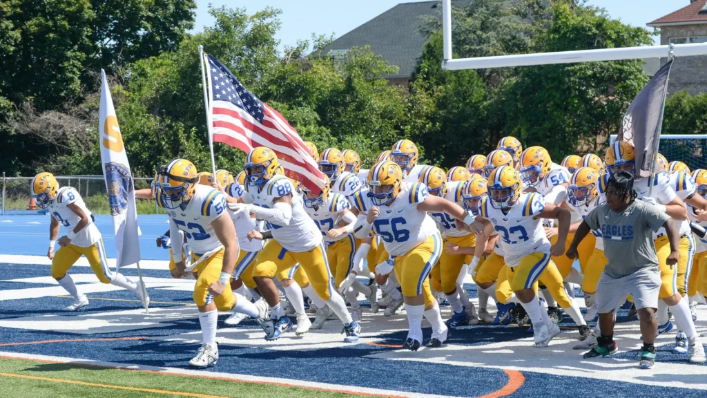 The St. Peter's Eagles football team charges onto the field in white and gold uniforms, carrying an American flag and team banners before a game.