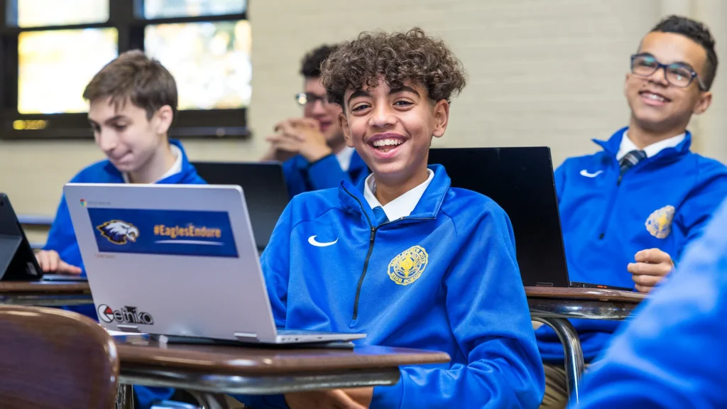 Students in blue school uniforms smile and work on laptops in a classroom, with one student in the foreground grinning broadly at the camera.