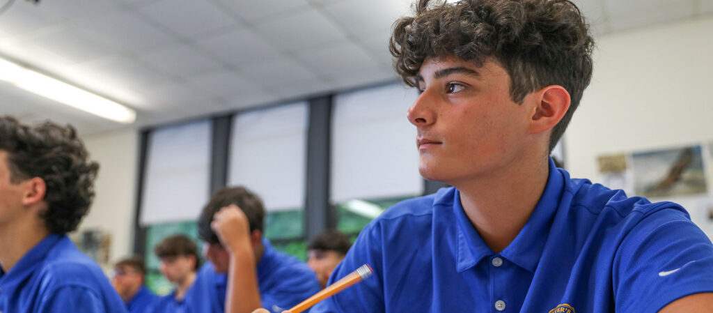 Student in a blue polo shirt attentively listening during class, holding a pencil, with classmates visible in the background.